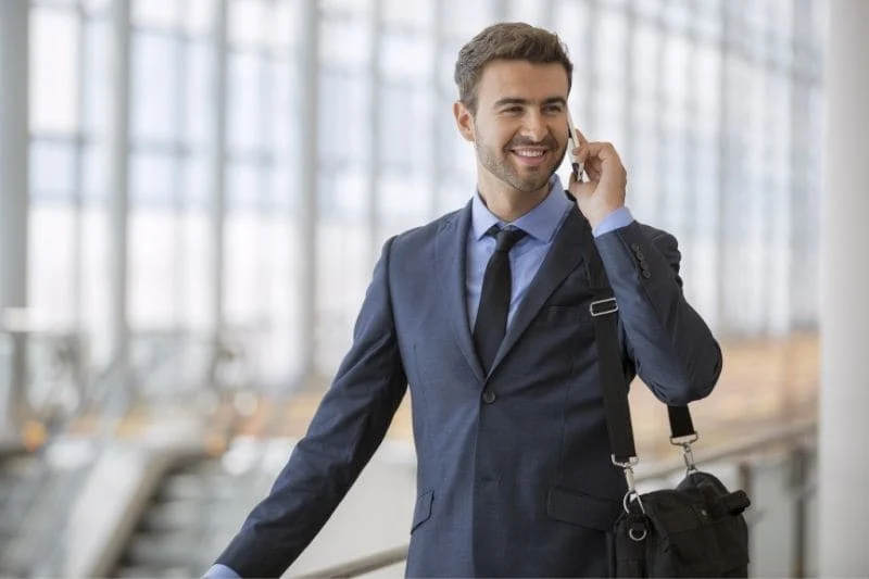 Un hombre en traje, habla por teléfono móvil, sonriendo, mientras lleva un maletín en un aeropuerto